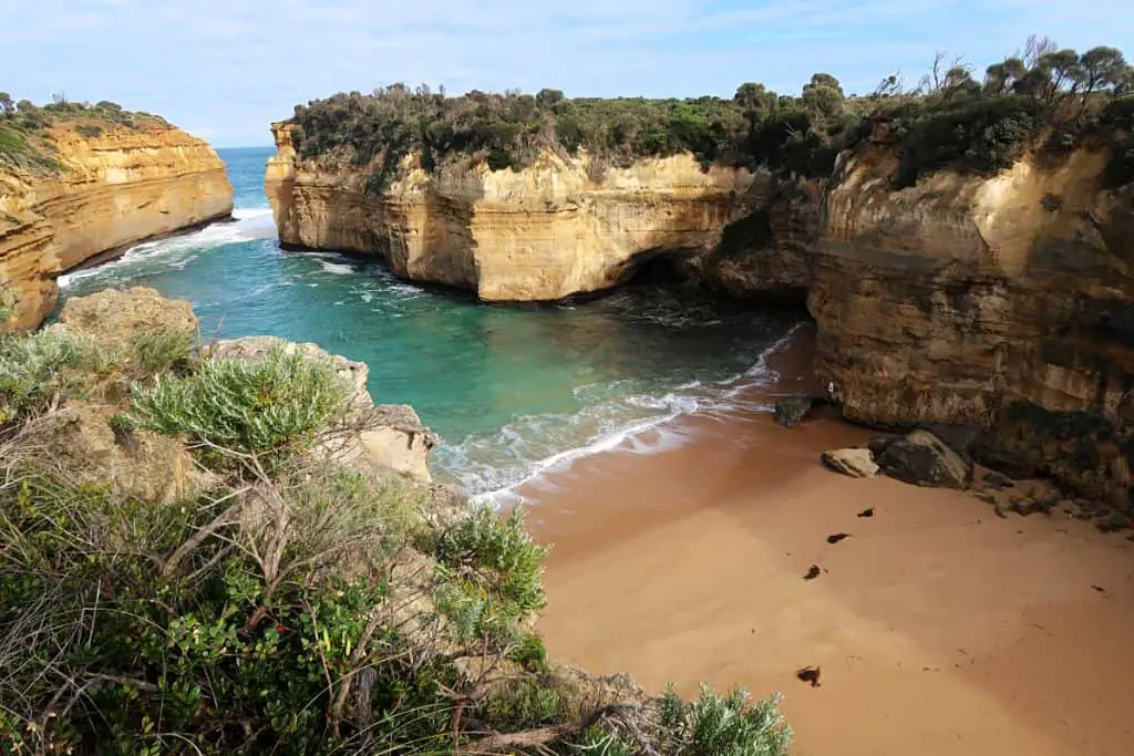 View of the golden sand at Loch Ard Gorge beach on the Great Ocean Road with giant cliffs, coastal greenery, and emerald water.