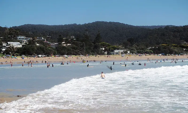 People swimming amongst the white capped waves at Lorne Beach on the Great Ocean Road in Victoria. The golden sand beach and green hills can be seen in the background.