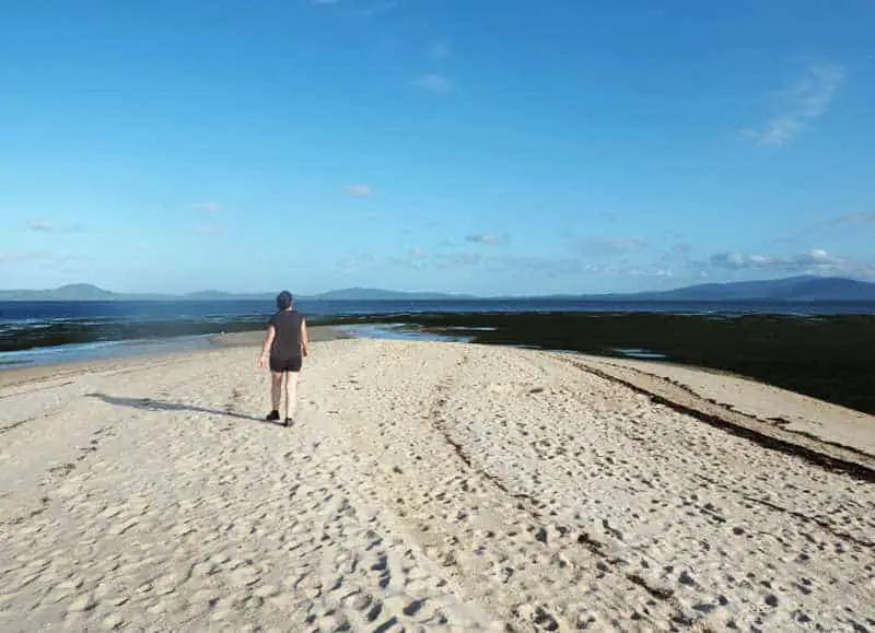 Woman walking along a Wilsons Prom Beach with bright blue sky and mountains in the distance.