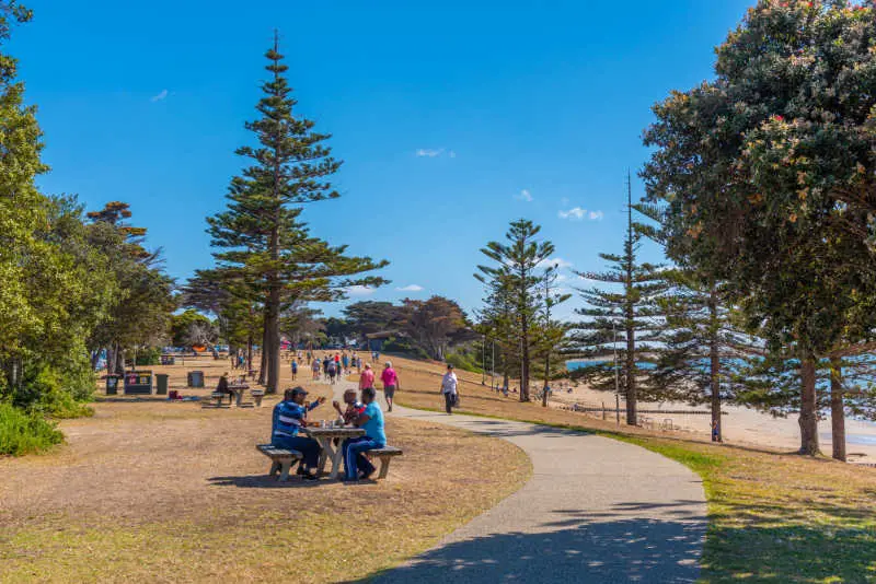 People sitting at picnic tables and waling at Torquay a Great Ocean Road town.  With blue sky and trees on a sunny day.