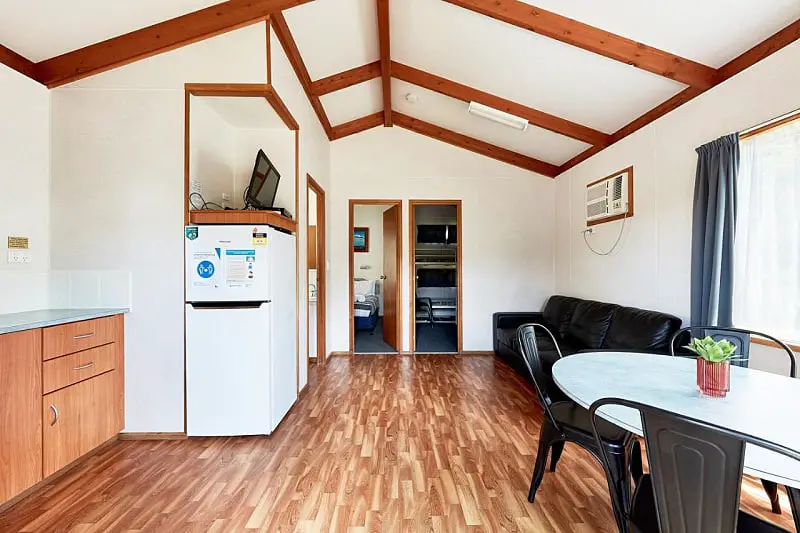 Open plan living, kitchen, and dining area at Kennett River Caravan Park on the Great Ocean Road in Victoria. There is a black vinyl couch and an oval table with black chairs and a small potted plant sitting on it. 