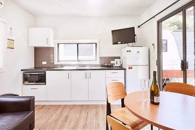 Open plan living, dining, and kitchen at a Lorne Foreshore Caravan Park cabin. There is a bottle of wine and two glasses sitting on the dining table.