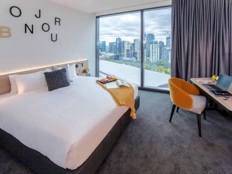 Guest room with floor to ceiling windows, bed, and desk at Novotel Melbourne South Wharf.