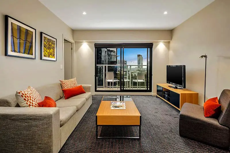 Seating area at a Quest Docklands guest room with couches, coffee table, television, and city view from floor to ceiling windows.