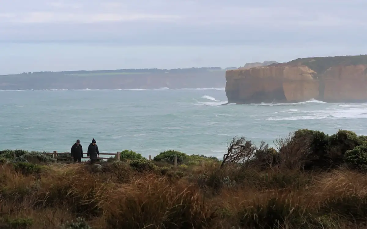 People walking along a trail on their Great Ocean Road 3 day itinerary with waves crashing into the cliffs in the background.