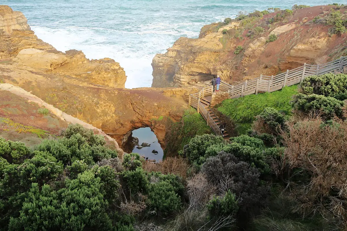 View of the Grotto a must-visit Great Ocean Road destination. With see-through archway, ocean, greenery, and people descending the wooden stairs down to the bottom of the cliff.