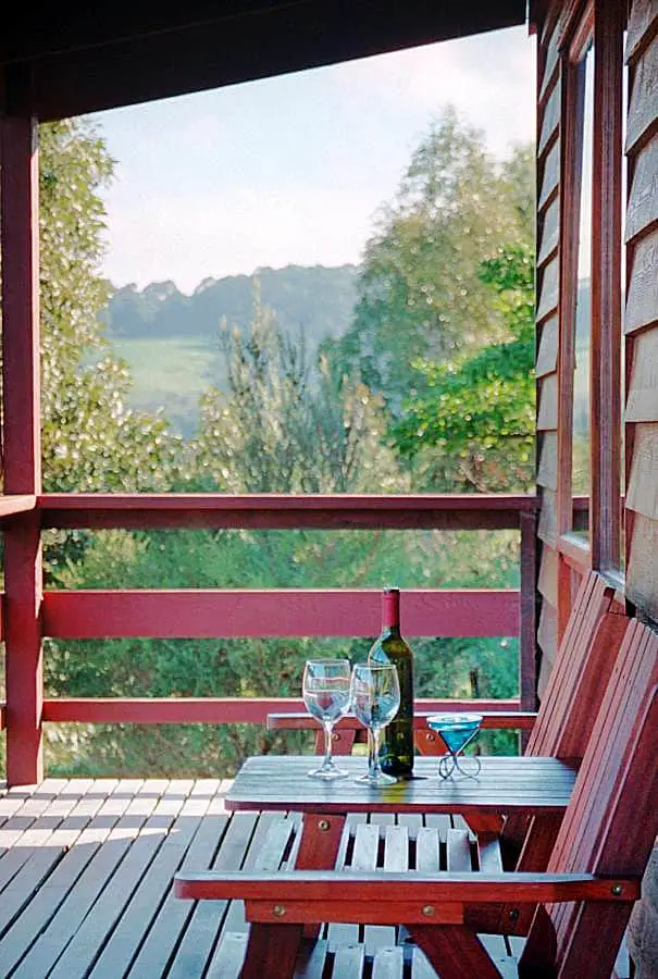 Wooden deck at Johanna River Farm & Cottages in the Otways with a table and chairs, a bottle of wine, wine glasses, and bush views.  