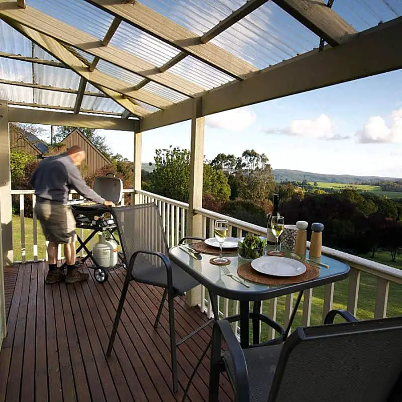 Verandah with views of the surrounding landscape, a person cooking on a BBQ and table set for dinner with wine glasses