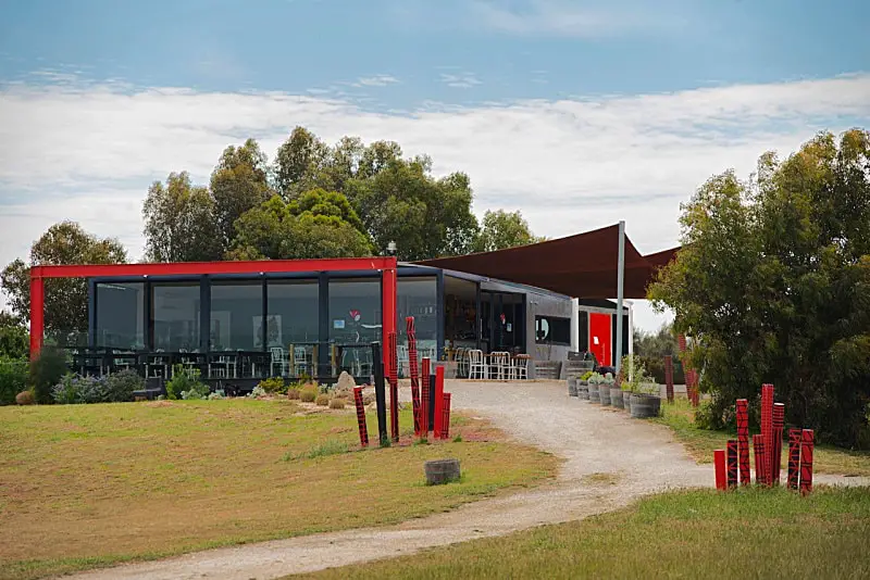 Banks Road Vineyard a Bellarine Peninsula winery with bright red features a dirt walking track and grass, trees, and blue sky with clouds. 