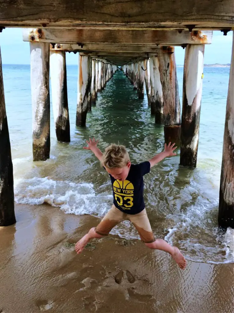 Child wearing wet shorts and a t shirt jumping under the Point Lonsdale Pier making a star shape  with the ocean behind him. 