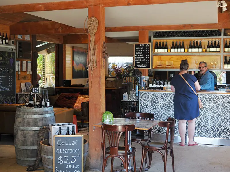 Woman buying wine from a winemaker at Curlewis Winery on The Bellarine. The cellar door has wood features and beams, wine barrels, table and chairs and a sign advertising discount wine for sale at $25.00.