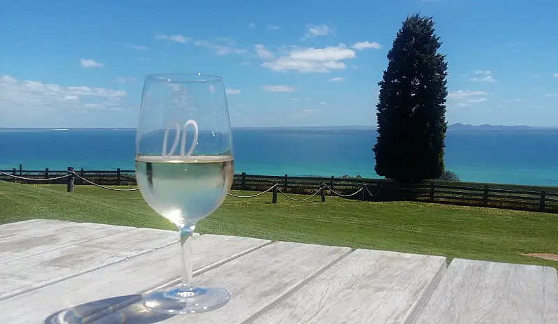 A glass of wine sitting on a table with views of the bay and a pine tree in the background at Jack Rabbit Winery on the Bellarine Peninsula in the Australian state of Victoria.
