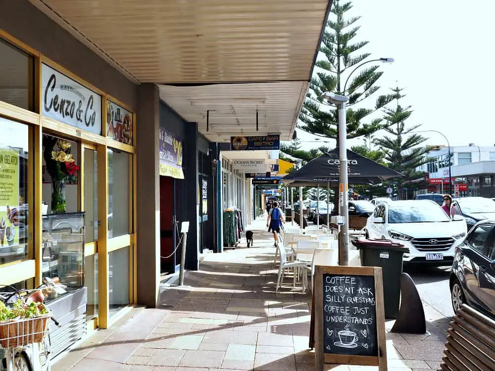 View of a street in Ocean Grove Bellarine Peninsula with a coffee sign, person walking the street, and trees.