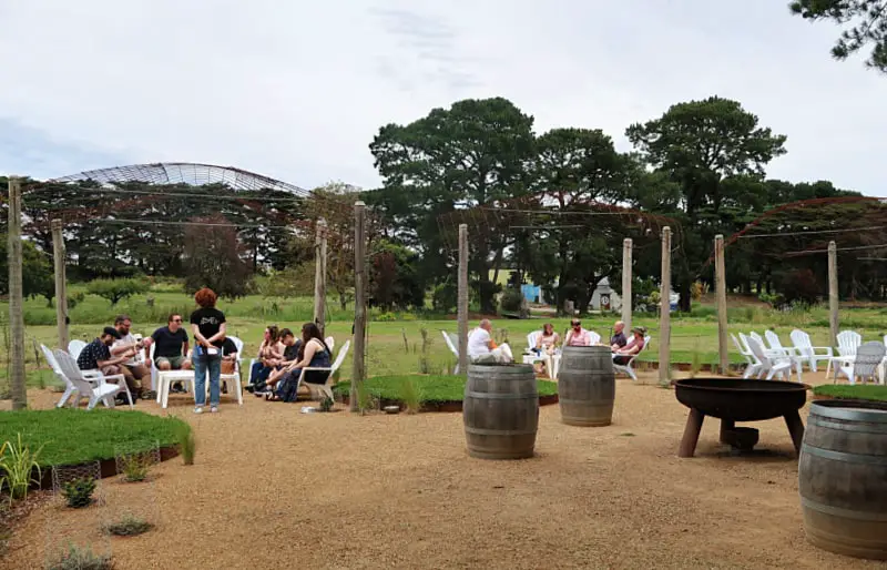 People relaxing outside on low chairs at The Whiskery with old wine barrels and a firepit in the outside area and trees in the background.