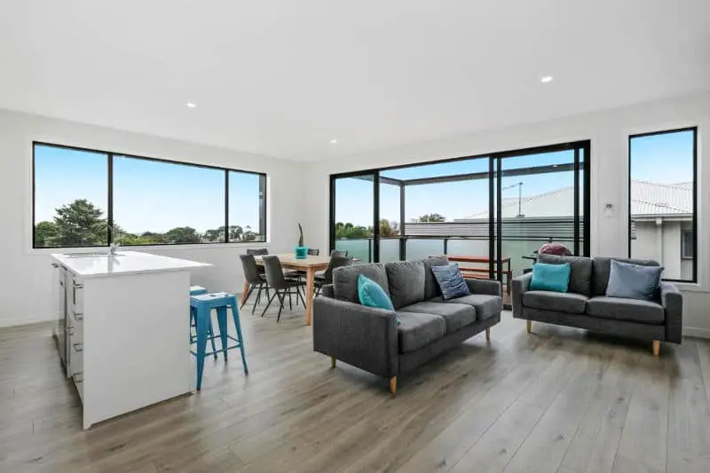 Living room at Waterline Property accommodation in Portarlington on the Bellarine Peninsula in Victoria with couches, a kitchen, dining table and large windows with views of the sky and trees.