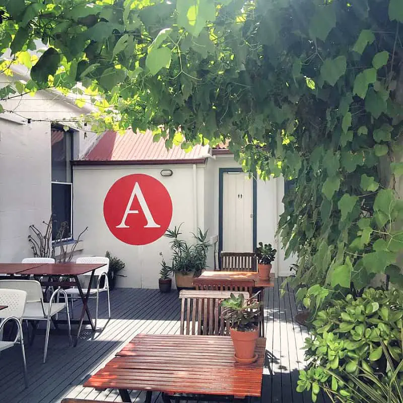 Tables and chairs in a courtyard at Athelstane Guesthouse in Queenscliff Victoria. A white letter A inside a red circle is painted on a white wall and the courtyard is shaded by a green vine. Athelstane House offers the best Queenscliff accommodation.