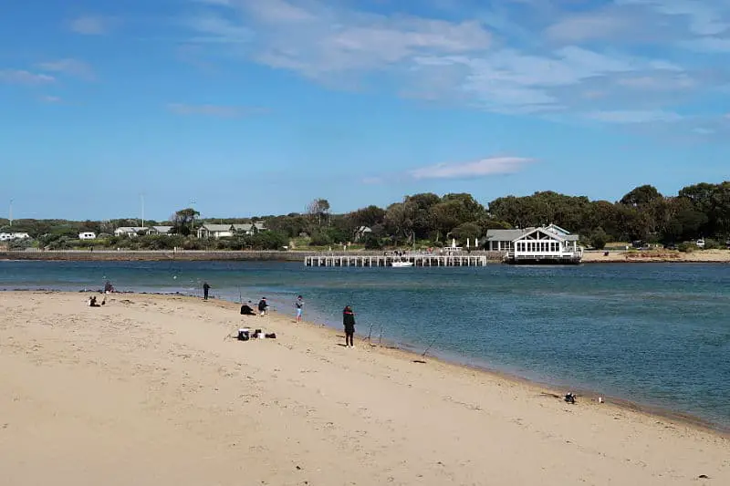 People fishing on the Barwon River beach in Ocean Grove with a view of At The Heads restaurant in Barwon Heads on the other side of the river.