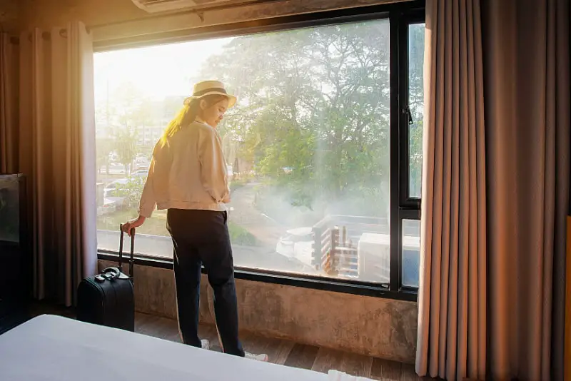 Woman standing at a large window admiring the view with her suitcase beside her. The best Bellarine Peninsula accommodation has views of the ocean and countryside.