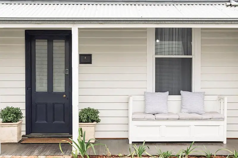 The entrance of Benambra Cottage featuring a sleek black door flanked by two potted plants, with a white wooden bench adorned with striped pillows to the right, all set against a cream exterior under a metal roof. Cute Queenscliff accommodation for couples.