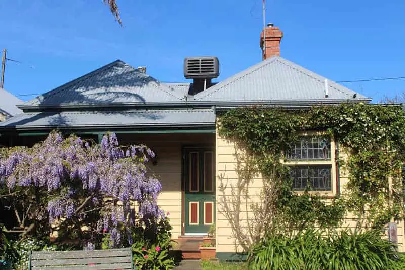 The cream coloured Benambra Guesthouse accommodation at Queenscliff in Victoria. The front entrance has a green door. There is a flowering lilac bush in the front garden and a green vine growing over the window and wall.  