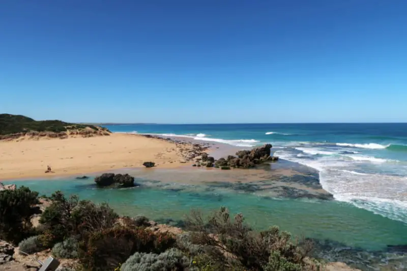View of the Blue Hole in Warrnambool with golden sand, blue white capped waves, rocks, coastal greenery and the aquamarine blue hole river mouth.