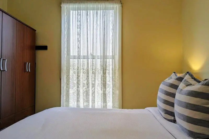 Cozy and well-appointed Learmonth Guesthouse bedroom in Queenscliff with a comfortable bed adorned with striped pillows, a wooden wardrobe, and a window dressed with elegant lace curtains.