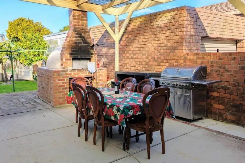 A quaint outdoor dining area at Learmonth Guesthouse featuring a wooden table set with floral tablecloth, chairs, and a brick barbecue setup under a pergola, with a green lawn and tree in the background. Perfect for guests to enjoy a sunny alfresco meal.