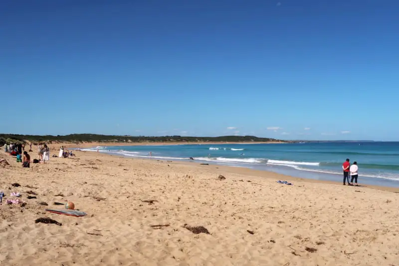 View of the coastline at McGennans Beach in Warrnambool> There are footprints in the sand and people enjoying the beach. McGennans is one of the popular beaches in Warrnambool.