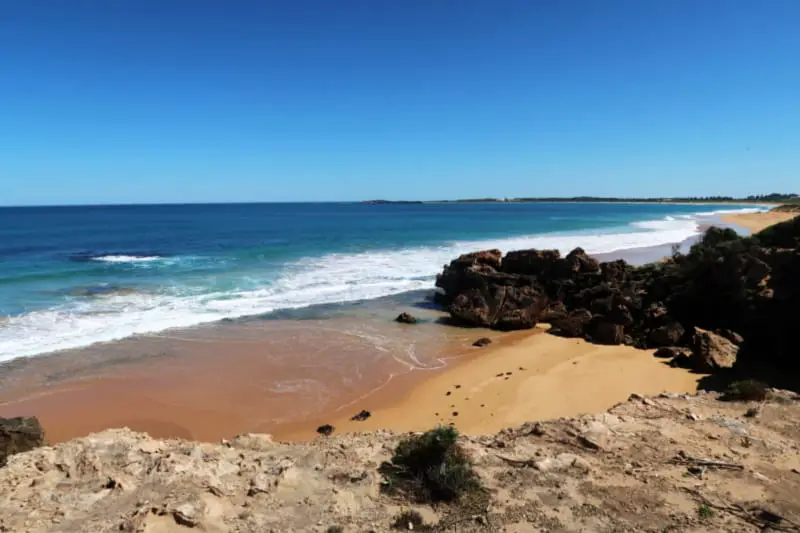 Moyjil Beach in Warrnambool with golden sands, white capped aquamarine waves and rocky outcrop. One of the many beautiful beaches in Warrnambool to visit.