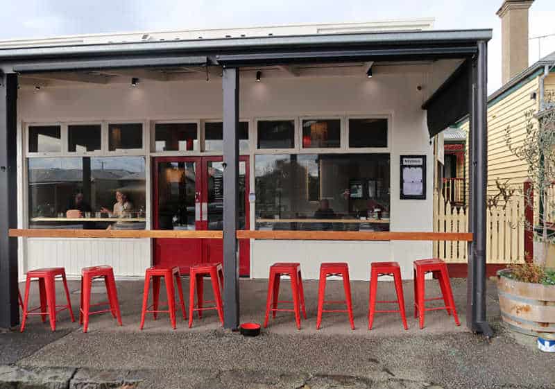 Outside eating area with a bench and red plastic stools at Nivana Queenscliff. There are people eating inside at the windows.