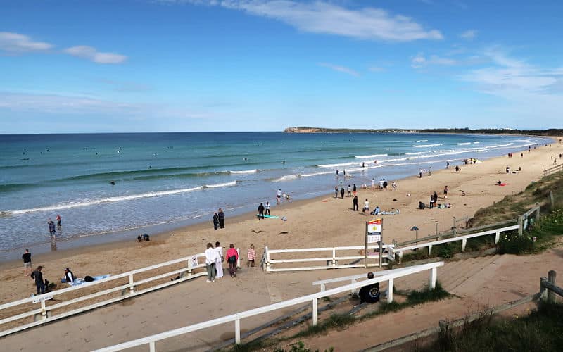 People enjoying the beach at Ocean Grove. There is a long stretch of sand and gentle white capped waves. The walkway leading down to the beach has a white fence and there are people admiring the beach. Visiting the beach is one of the best things to do in Ocean Grove on the Bellarine Peninsula.