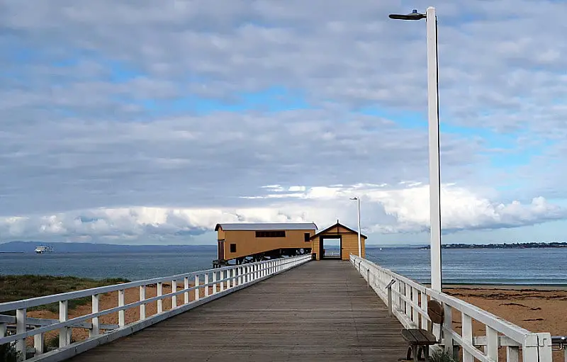View of the Queenscliff Pier with both the Shelter Shed and Boat Shed at the end of the jetty. There is a tall light attached to the white balustrade, and cloudy blue skies. Fishing and walking along the pier are popular activities in Queenscliff Victoria.