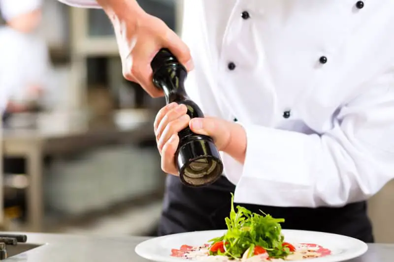 Chef in a white coat using a pepper grinder to season a green salad on a white plate. There are many Queenscliff restaurants where you can enjoy a meal in this seaside village on the Bellarine Peninsula in Victoria.