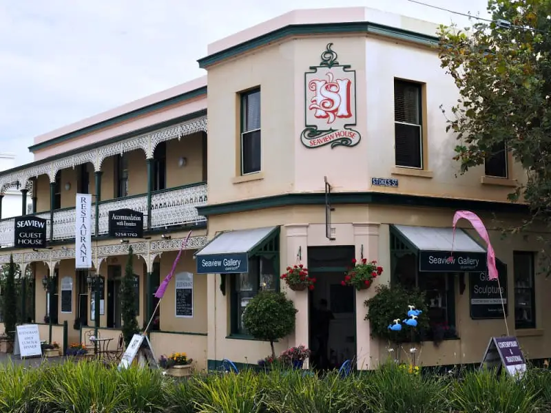 HIstorical cream coloured corner building with window awnings and hanging baskets with red flowers that houses Seaview Gallery one of the best art galleries in Queenscliff.