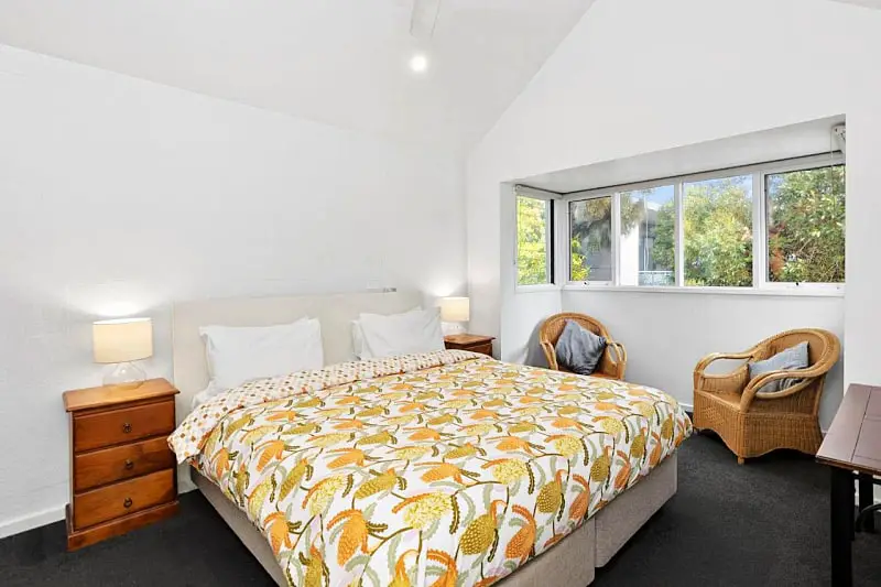 Guest room at Terrace Lofts Ocean Grove accommodation. There is a double bed with a bright yellow doona cover, two cane chairs, side tables and greenery can be seen through the window.