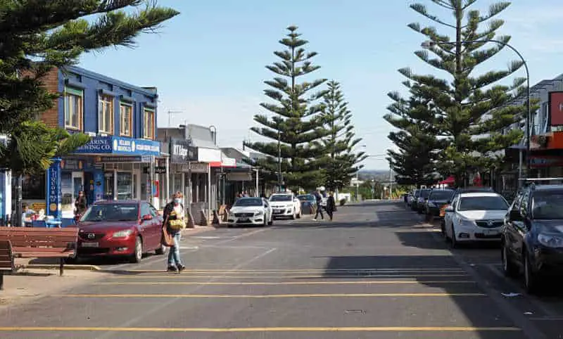 Shopper crossing the road in Ocean Grove Victoria. There are parked cars and large pine trees lining the road. Shopping for surf gear is one of the popular things to do in Ocean Grove.