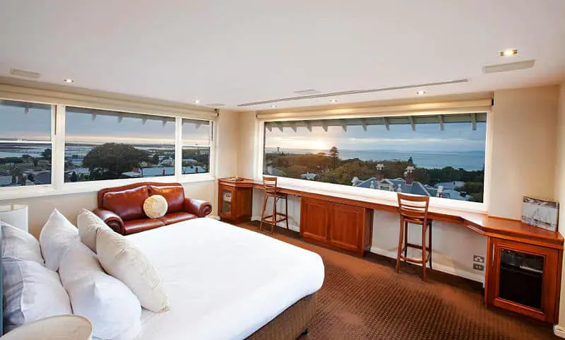 Guest room at the Vue Grand Hotel in Queenscliff on the Bellarine Peninsula. There is a bed, a leather couch, two rectangle windows with views over the town to the ocean, and a desk along one wall under a window.   