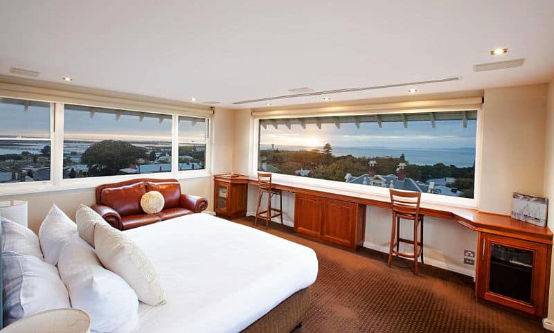 Guest room at the Vue Grand Hotel in Queenscliff on the Bellarine Peninsula. There is a bed, a leather couch, two rectangle windows with views over the town to the ocean, and a desk along one wall under a window.