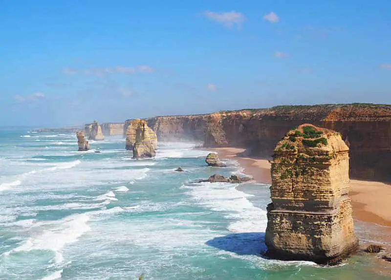 The Twelve Apostles giant stone stacks on a clear day with fluffy white clouds and gentle white capped waves. Seeing the 12 Apostles is included in all the best Great Ocean Road 2 days tours from Melbourne.