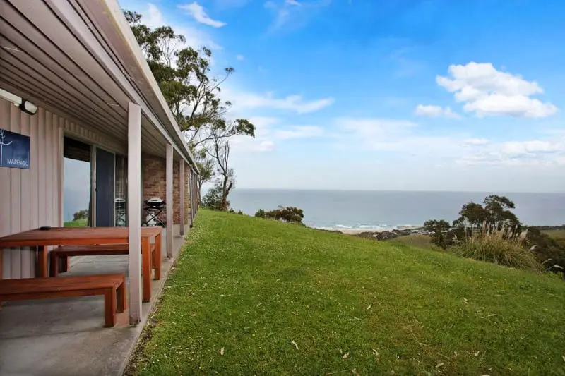 Decked area with barbecue and outdoor furniture next to a large grassy area with ocean views.