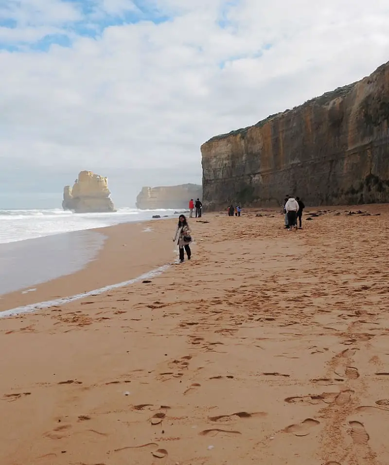 People walking on the beach below the cliffs on the Great Ocean Road. A rock stack rising out of the ocean can be seen in the background.