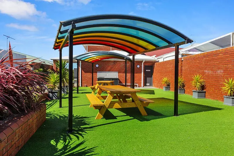 Outdoor area with picnic tables and a colourful canopy cover, artificial turf and potted palms.
