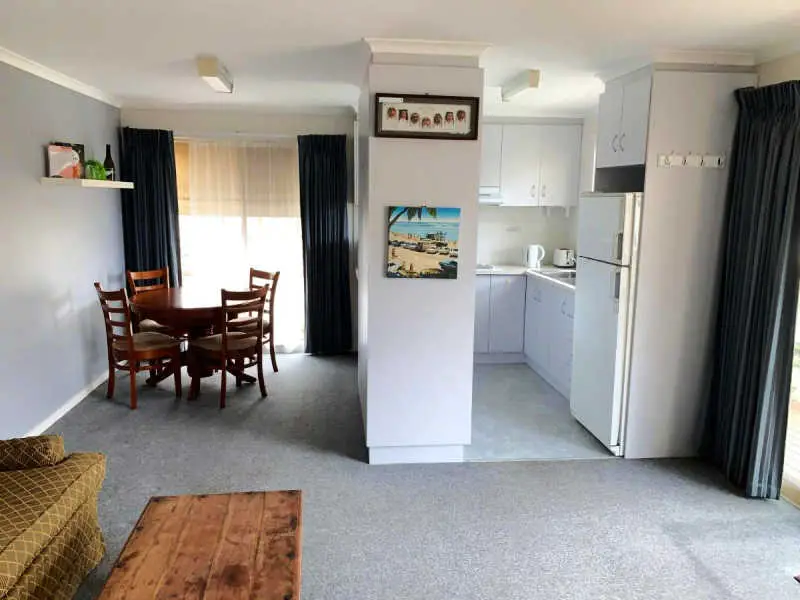 View of the white galley kitchen and meals area at the Portarlington Holiday Apartment. There is a round table with chairs and pictures on the wall.