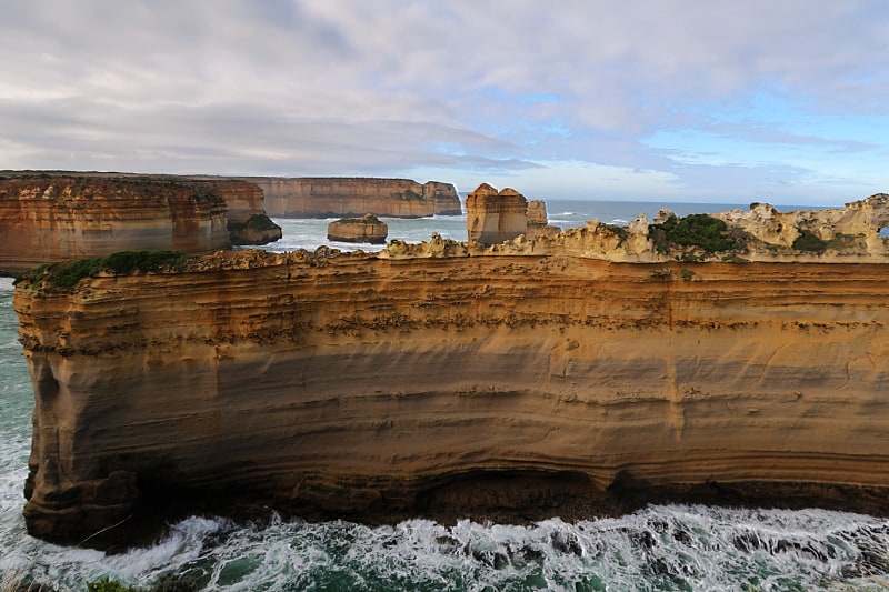 The Razorback rock formation on the Great Ocean Road. There are waves crashing against its base and views of other rock formations in the background. The Razorback can be seen on one of the tours of the Great Ocean Road from Melbourne.