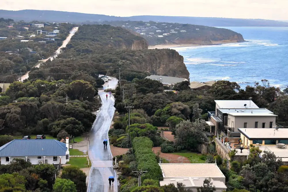 View of Aireys Inlet from the top of Split Point LIghthouse on a rainy day. There are people walking along the road under umbrellas. The ocean and coastline can be seen.