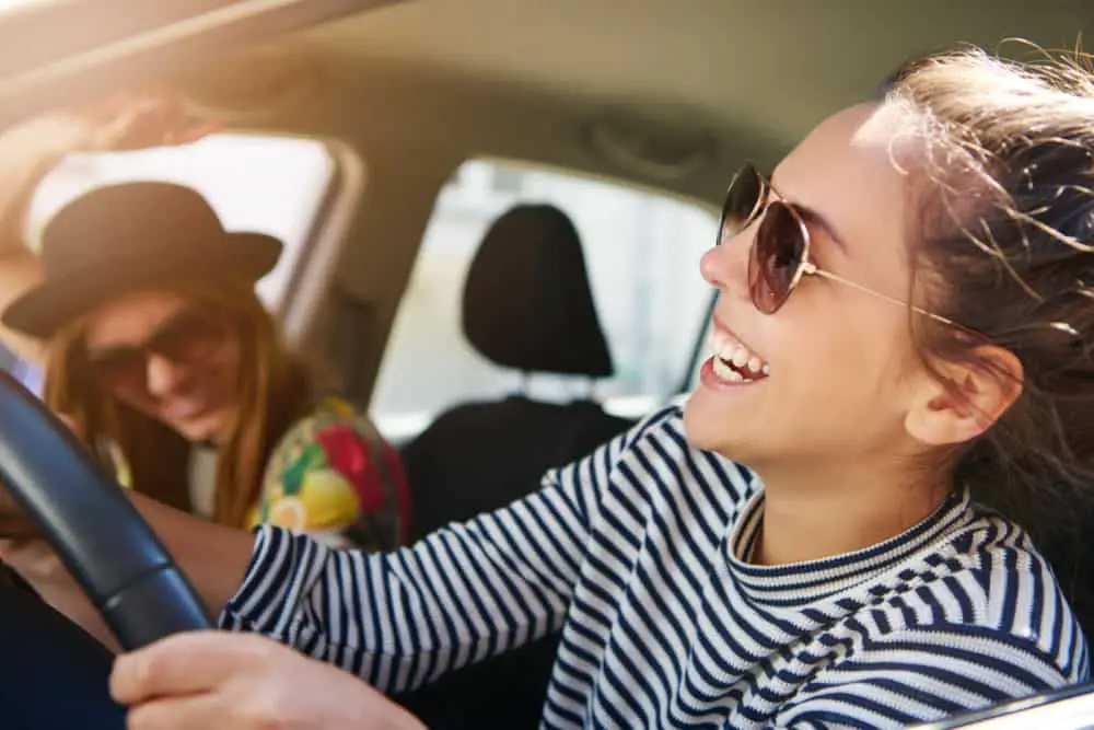 Two women taking a road trip. A woman is leaning out of the car window smiling. 