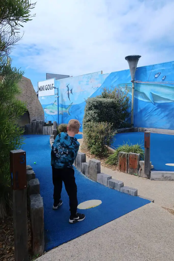 A small boy with blonde hair playing a shot at Great Ocean Road Mini Golf. A visit here is one of the fun things to do in Aireys Inlet with kids.