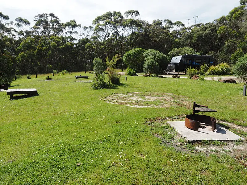 Hammonds Road Campground with grass campsites, firepits, picnic tables and a caravan parked amongst the trees. This is a popular spot for camping the Great Ocean Road. 