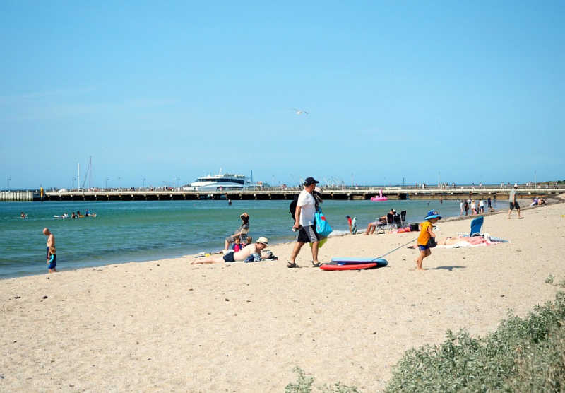 Man walking across the sand while others lie sunbaking at Portarlington Beach the pier and Docklands Ferry can be seen in the background. This calm swimming beach is a popular Portarlington attraction.