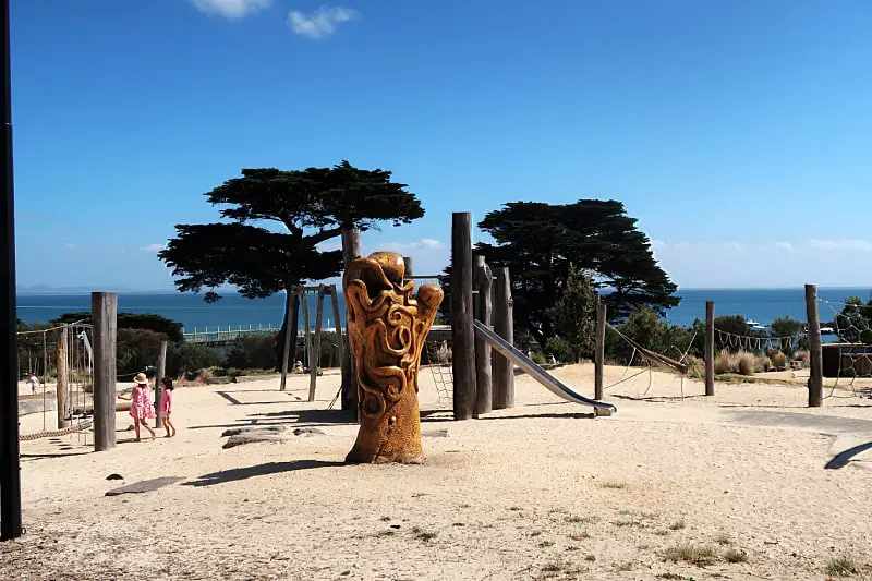Children play near a distinctive, carved wooden sculpture at a sandy park in Portarlington, with the serene ocean and clear blue sky in the background.